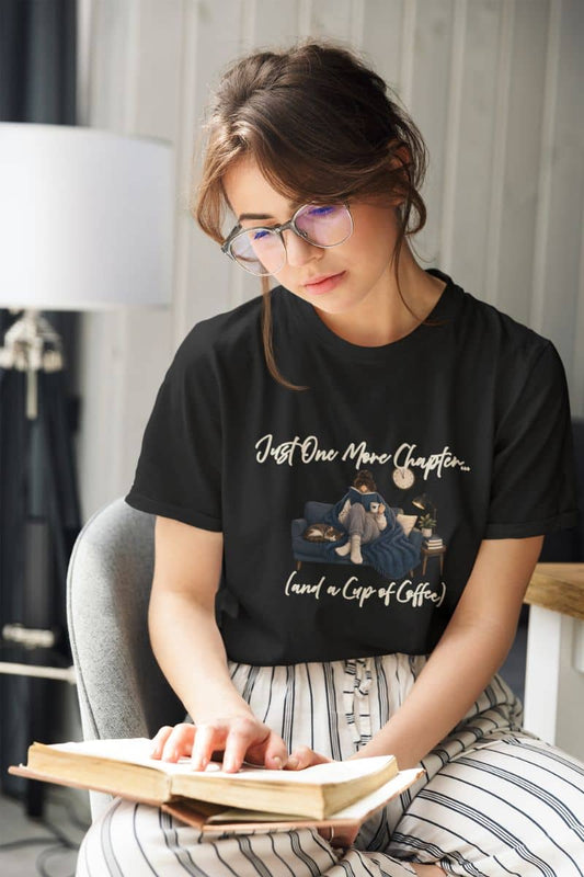 Book lover t shirt with a woman wearing a black graphic t-shirt featuring the text 'Just One More Chapter', reading a book in a cozy indoor setting. 