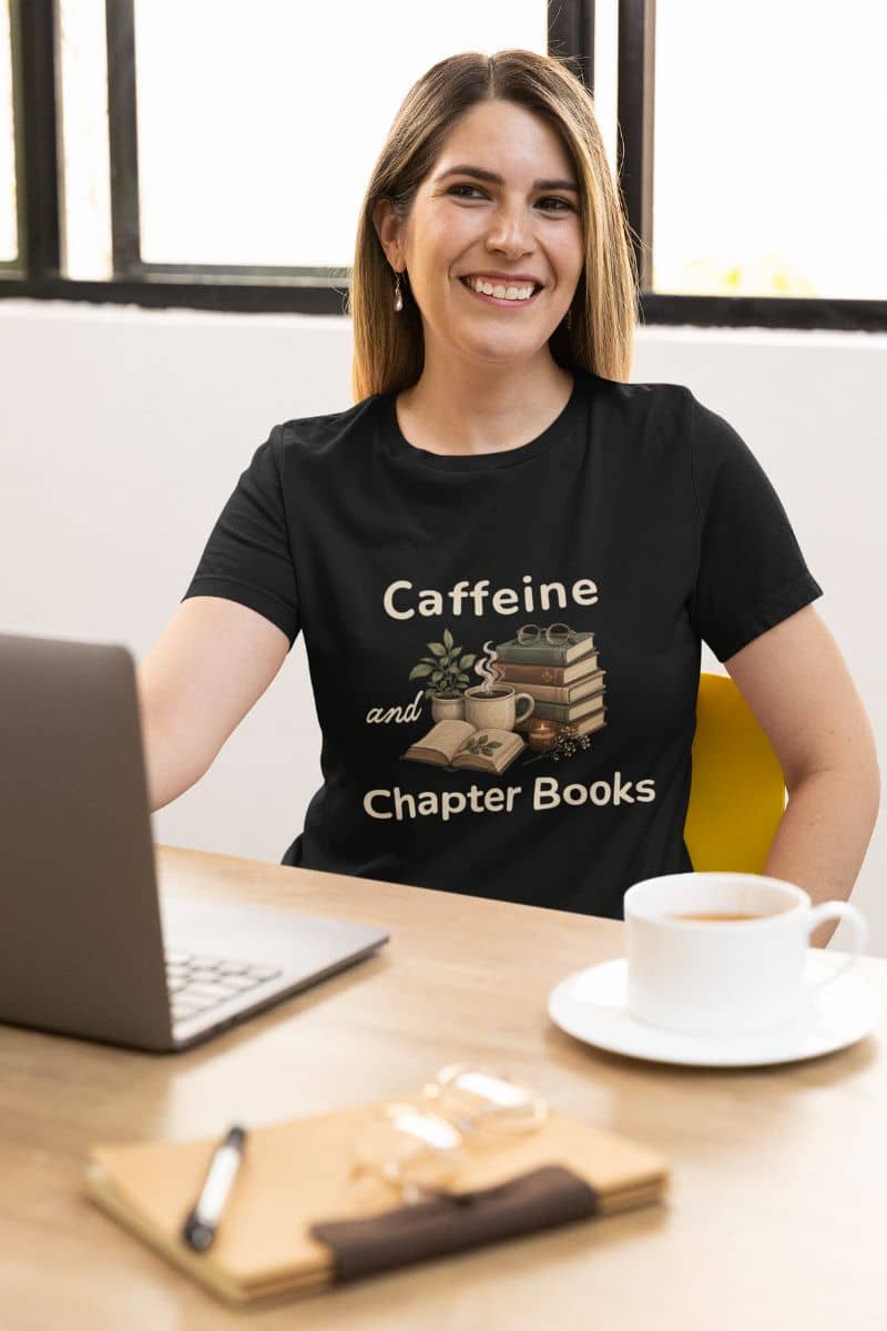 Book lover tees with a smiling woman sits at a desk wearing a black graphic T-shirt that reads 'Caffeine and Chapter Books' 