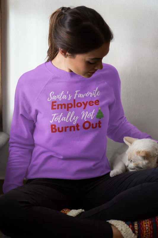 Burnout humor sweatshirt with a person wearing a purple sweatshirt with text 'Santa’s Favorite Employee Totally Not Burnt Out' sitting on a couch with a cat.