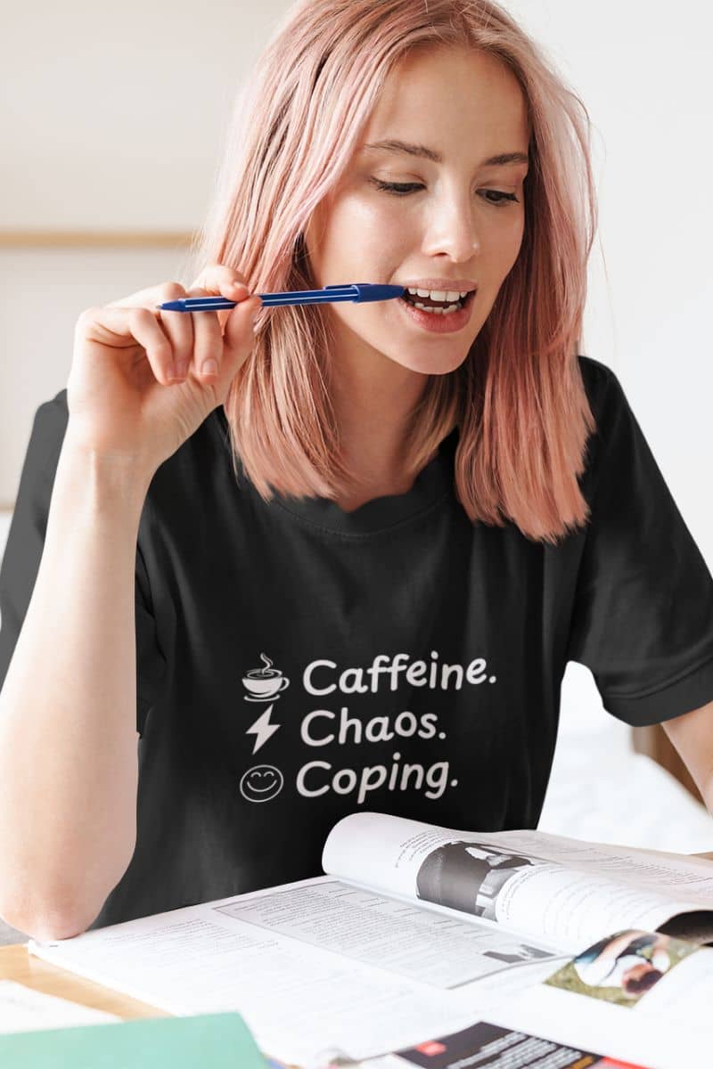 Caffeine humor tee with a person wearing a black t-shirt with 'Caffeine. Chaos. Coping.' text, holding a pen.