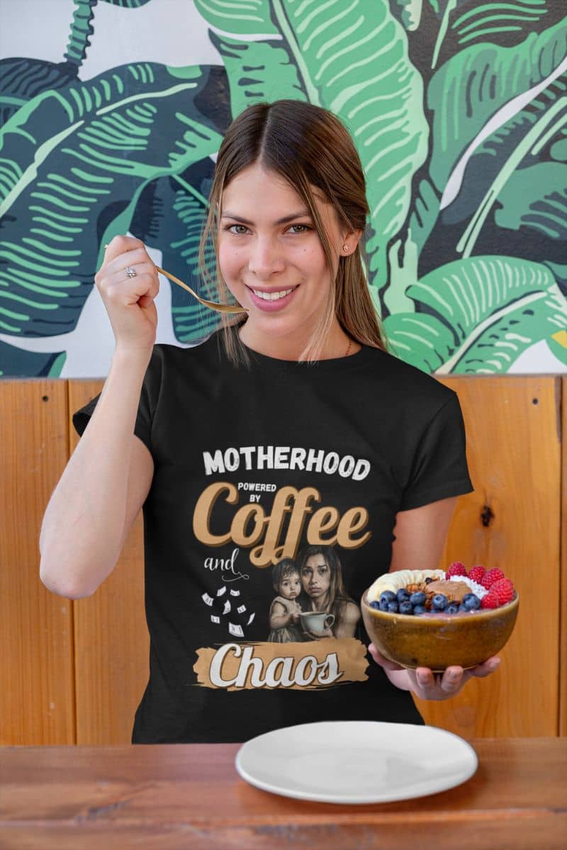 Chaos and coffee shirt with a woman wearing a black t-shirt with text 'Motherhood: Powered by Coffee and Chaos.', holding a bowl of fruit, in front of a green leafy mural.
