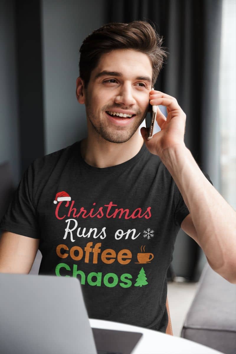 Christmas caffeine shirt with a man wearing a black t-shirt with text 'Christmas Runs on Coffee & Chaos', talking on a phone indoors.