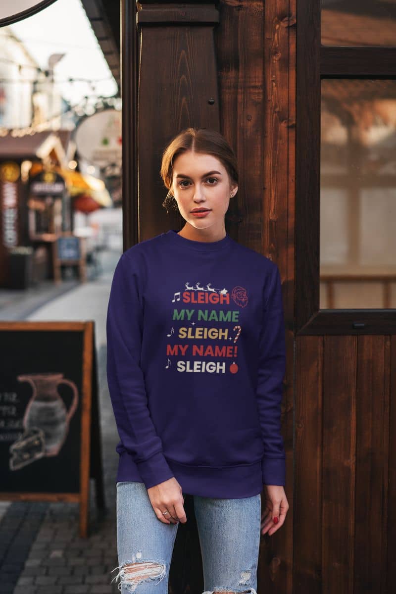 Christmas crewneck with a person wearing a purple sweater with text 'Sleigh My Name, Sleigh My Name' and graphics, standing in front of a wooden door.