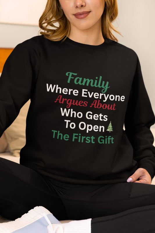 Christmas family humor sweatshirt with a person wearing a black sweatshirt with humorous text 'Family: Where Everyone Argues About Who Gets To Open The First Gift'