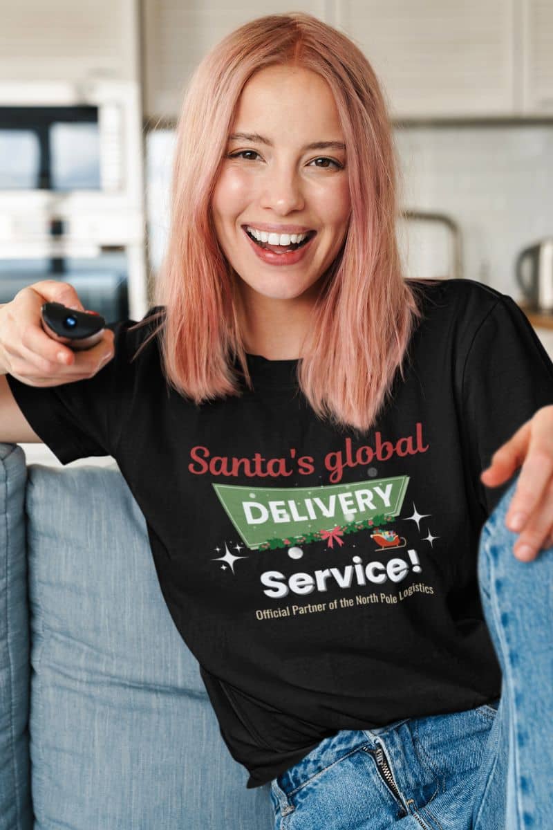 Christmas graphic tee with a person wearing a black t-shirt with 'Santa's global Delivery Service!' text, sitting on a couch.