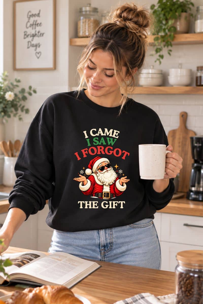Woman holding a white coffee mug in a cozy kitchen, wearing a black Christmas party sweatshirt with the funny “I Came. I Saw. I Forgot the Gift” Santa graphic.