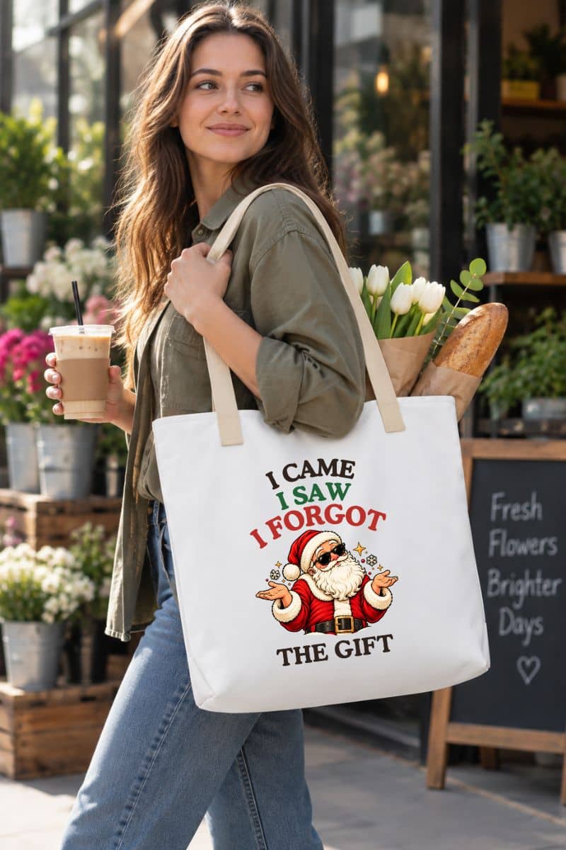 Woman carrying a Christmas tote bag with beige handles and “I Came. I Saw. I Forgot the Gift” Santa design while holding iced coffee outside a flower shop.
