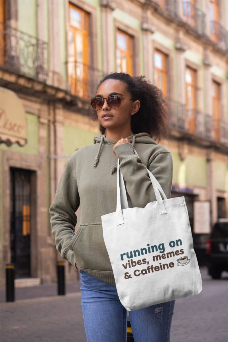 Coffee chaos carryall with a woman holding a tote bag with text 'running on vibes, memes & caffeine' on a city street