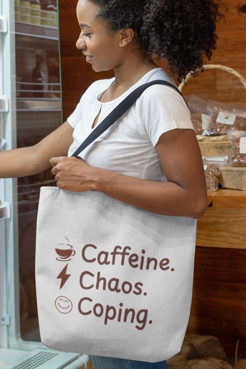 Coffee-fueled carryall with a woman holding a tote bag with 'Caffeine. Chaos. Coping.' text in a store setting.