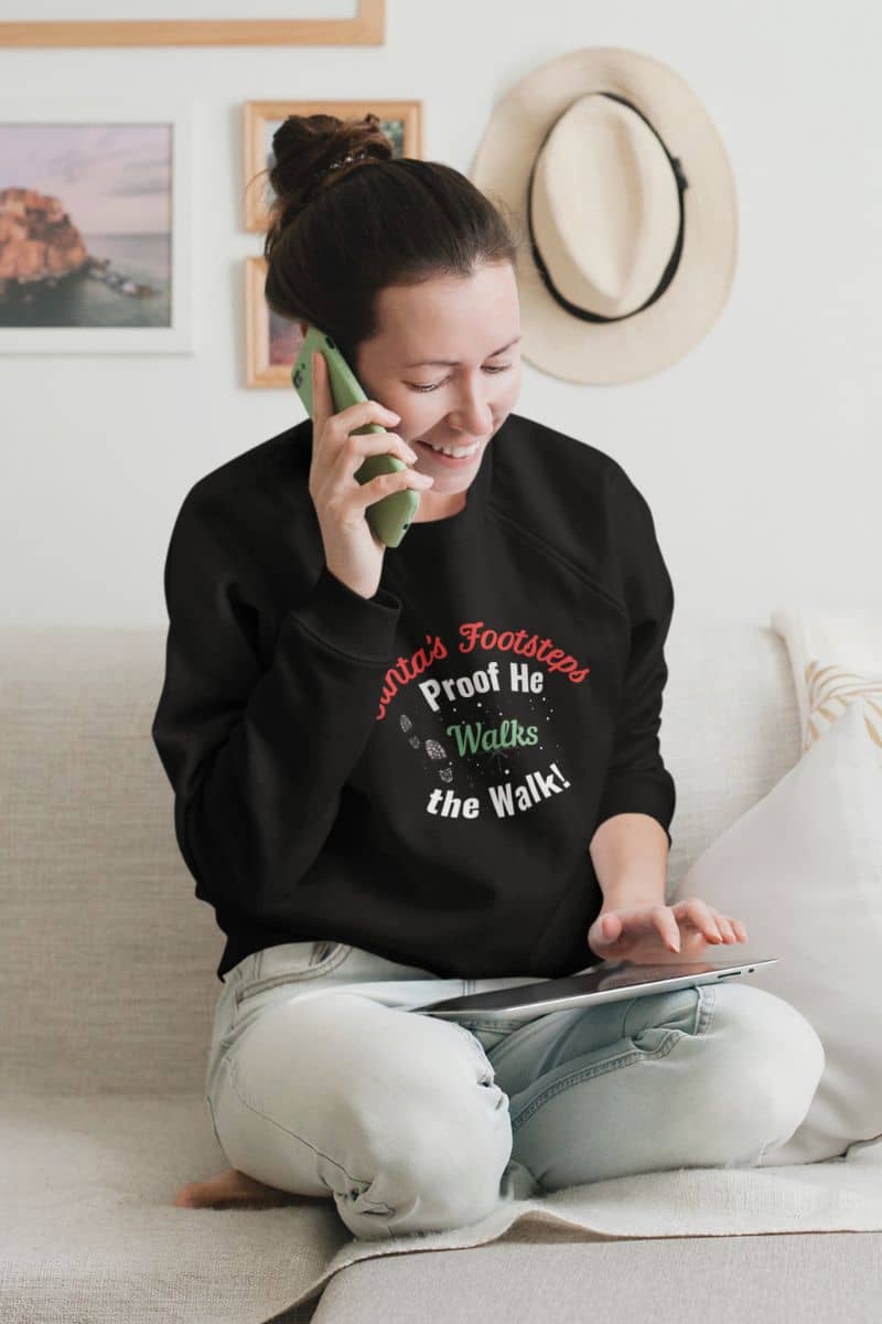 Cozy Santa sweatshirt with a woman sitting on a couch wearing a black sweatshirt with text 'Santa's Footsteps - Proof He Walks The Walk!', using a tablet and talking on a phone.