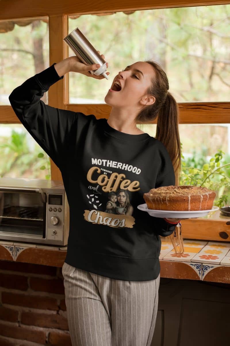 Cozy motherhood sweater with a woman in a kitchen holding a cake and a coffee cup, wearing a black sweatshirt with text 'Motherhood powered by Coffee and Chaos'.