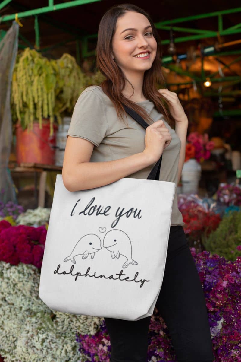 Cute dolphin tote bag with a woman holding a tote bag with 'I love you dolphinately' design in a market setting.