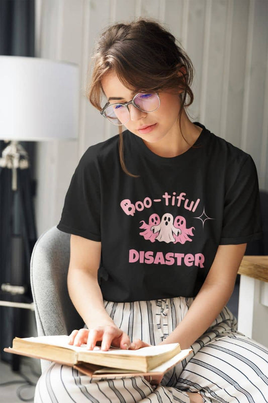 Cute halloween t-shirt with a woman wearing a black t-shirt with 'Boo-tiful Disaster' design, reading a book indoors.