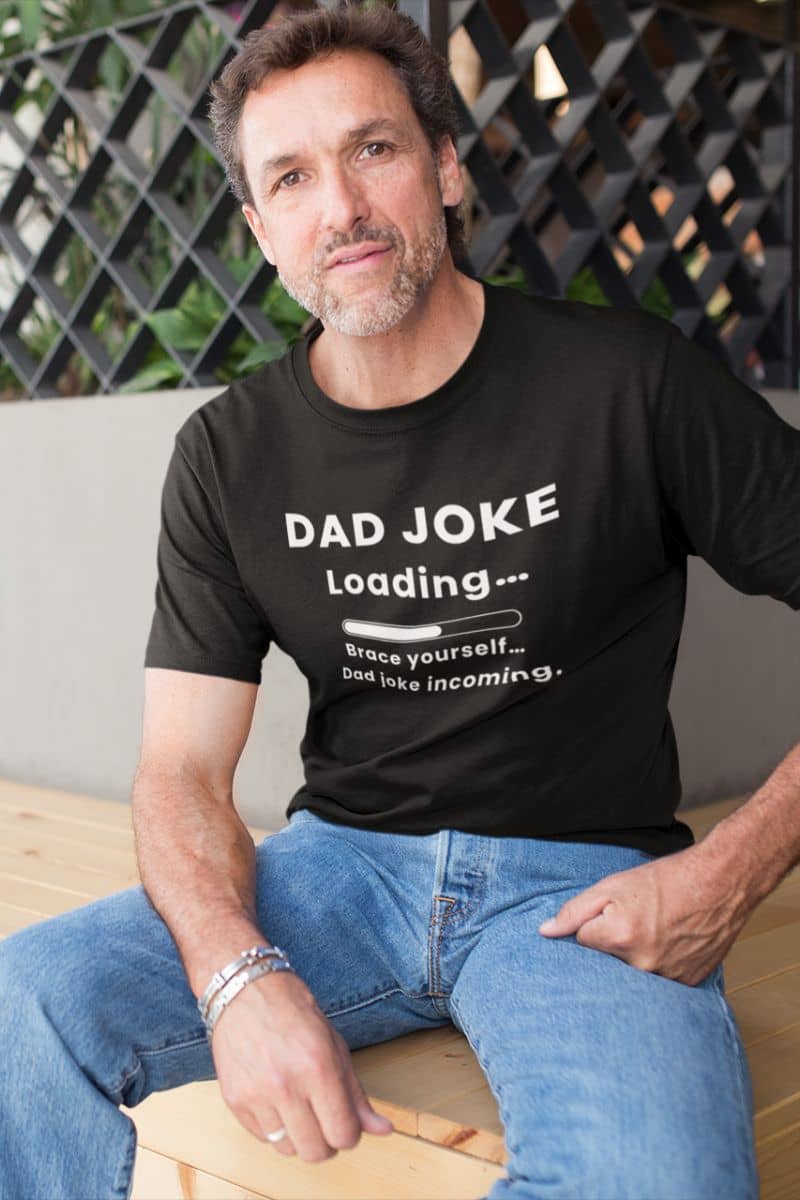 Dad joke shirt with a man wearing a black t-shirt with 'Dad Joke Loading, Brace yourself…Dad joke incoming.' text, sitting on a wooden bench.