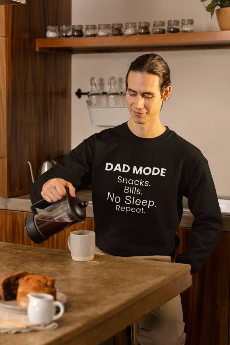 Dad life sweatshirt with a person pouring coffee wearing a black sweatshirt with text 'DAD MODE:  Snacks.  Bills. No Sleep. Repeat.', standing in a kitchen.