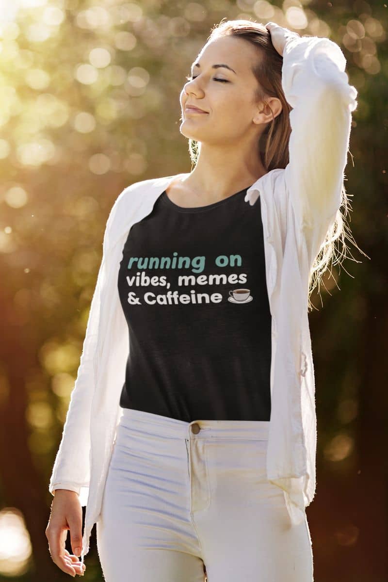 Daily fuel graphic t-shirt with a woman wearing a black t-shirt with text 'running on vibes, memes & caffeine' outdoors.