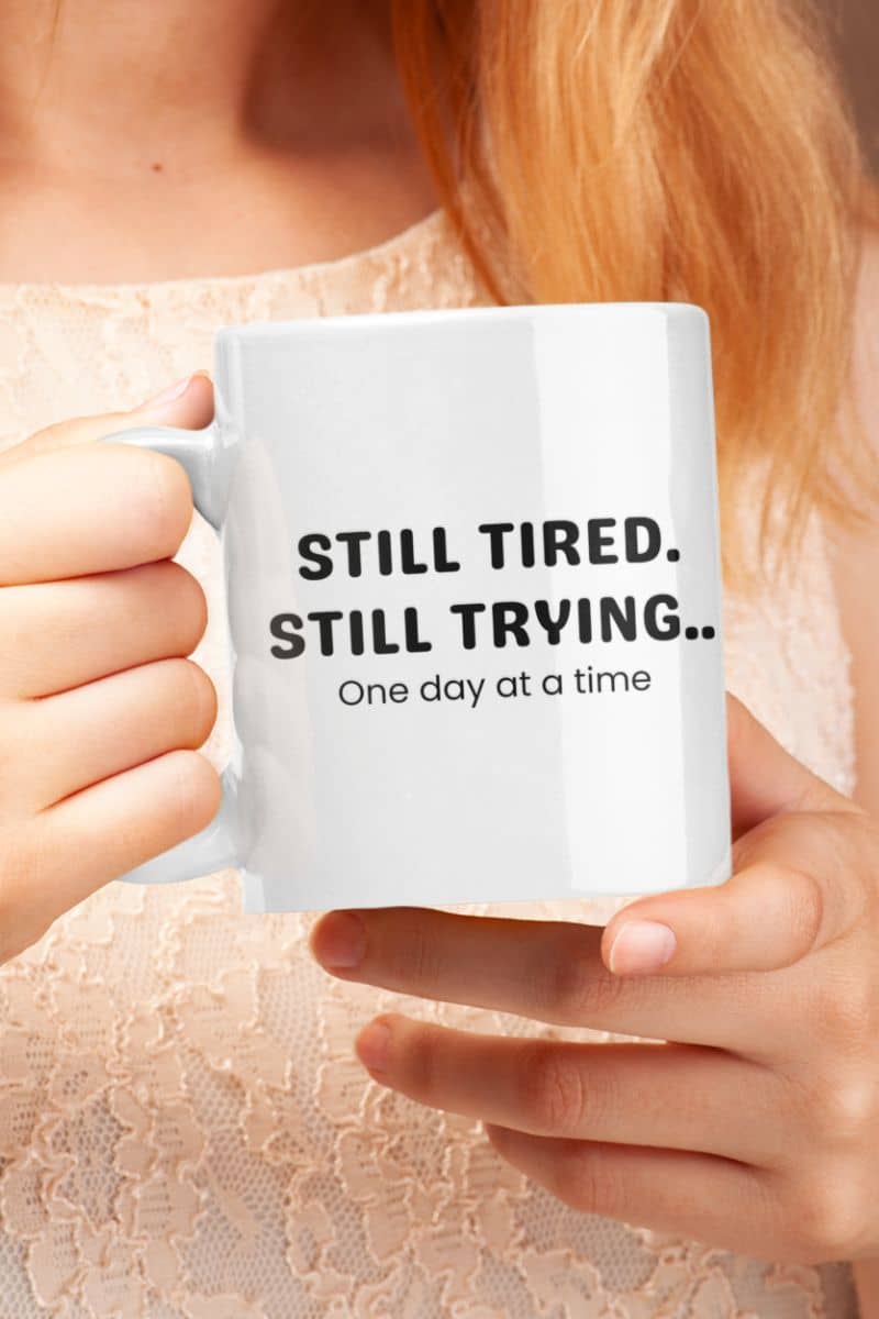 Daily motivation coffee mug with a person holding a mug with 'STILL TIRED. STILL TRYING.. One day at a time' text.
