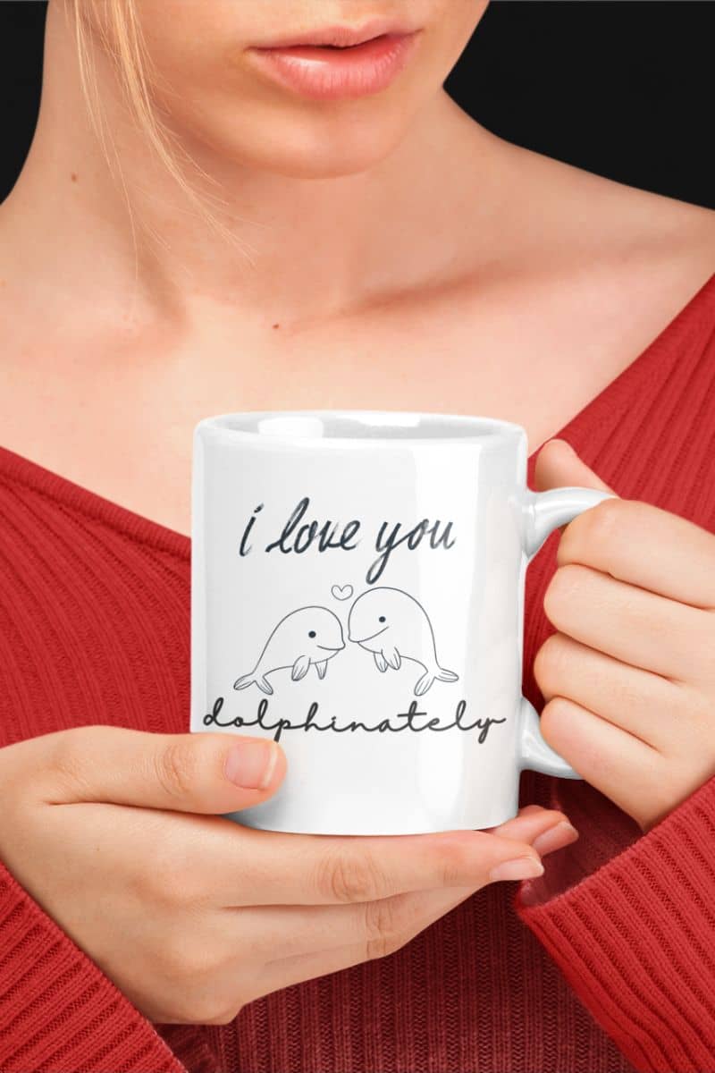Dolphin love coffee mug with a person holding a white mug with 'I love you dolphinately' text and dolphin illustrations.