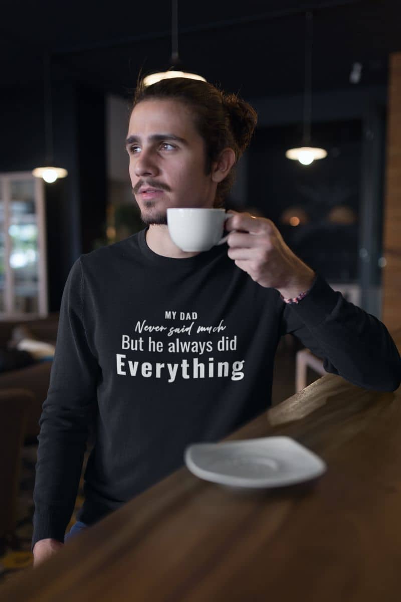 Emotional dad sweatshirt with a man holding a coffee cup in a dimly lit room, wearing a black sweatshirt with text 'My Dad Never Said Much, He Always Did Everything'.