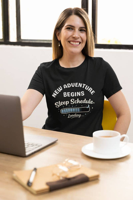 Exhausted parent t-shirt with a woman wearing a black t-shirt featuring the text 'New Adventure Begins, Sleep Schedule Loading…', sitting at a desk with a laptop and coffee.