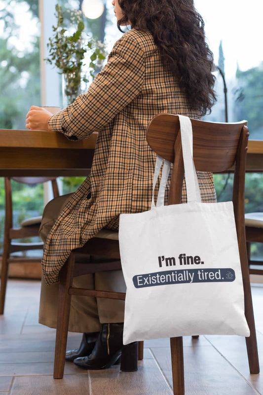 Existentially tired tote bag with a person sitting at a table with a tote bag that reads 'I’m fine. Just existentially tired'
