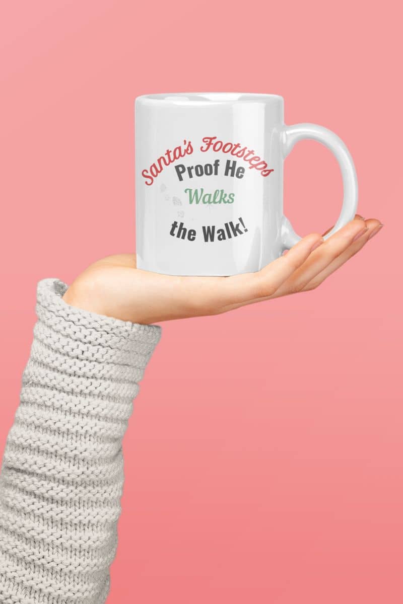 Festive holiday coffee mug with a mug with text 'Santa's Footsteps - Proof He Walks The Walk!' held by a person wearing a textured sleeve against a pink background