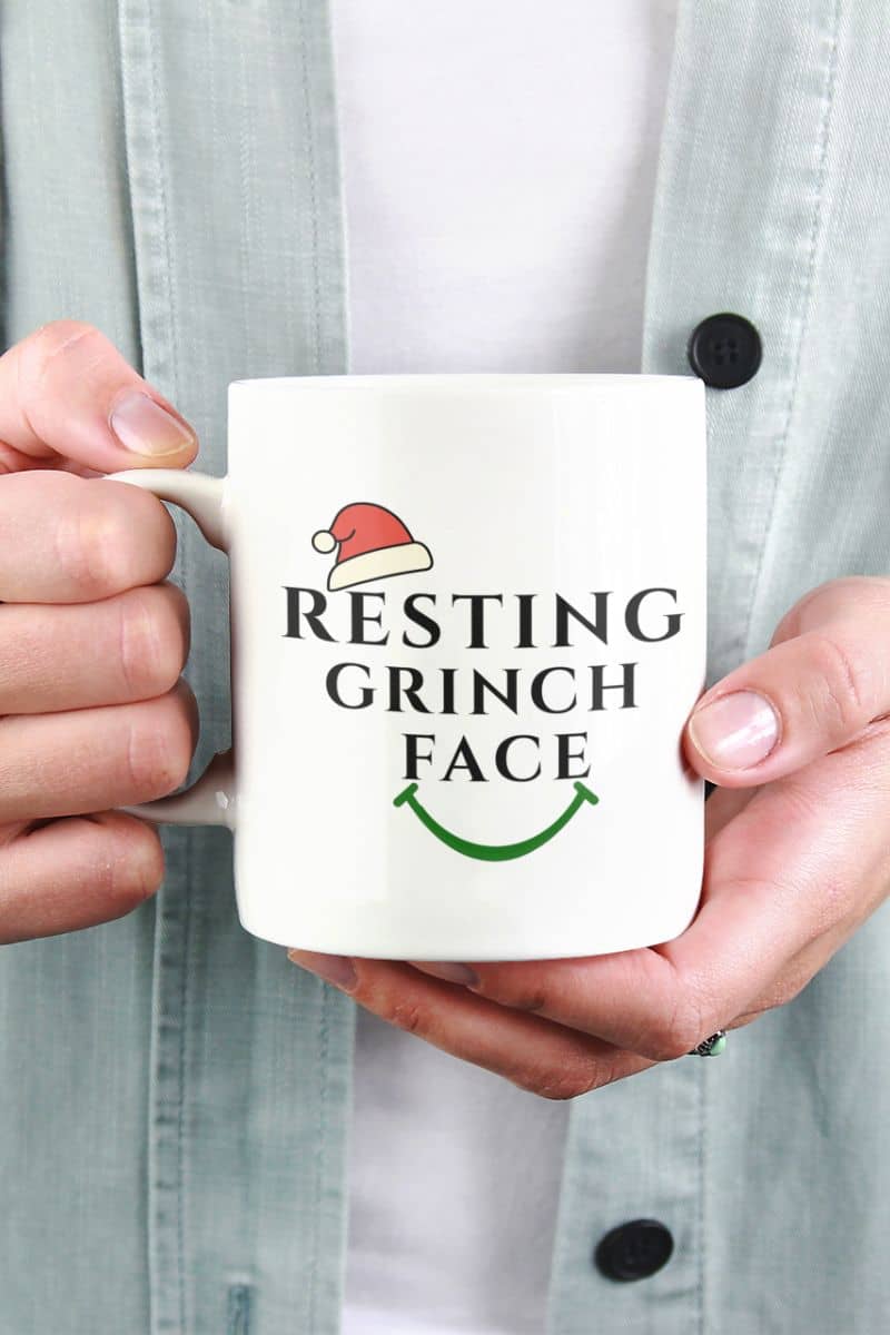 Festive office mug with a person holding a mug with 'Resting Grinch Face' text and Santa hat graphic.