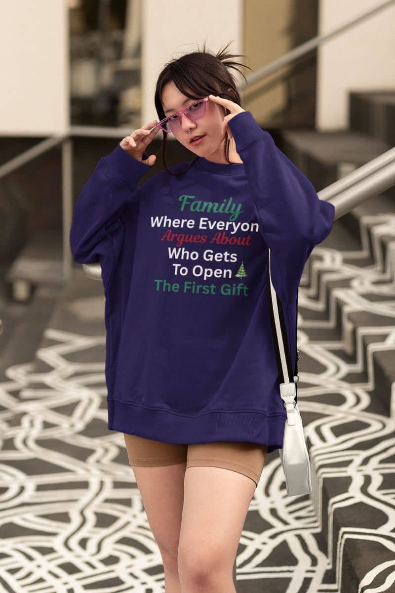 Festive quote sweatshirt with a person wearing a purple sweatshirt with text 'Family: Where Everyone Argues About Who Gets To Open The First Gift', standing on a patterned floor.