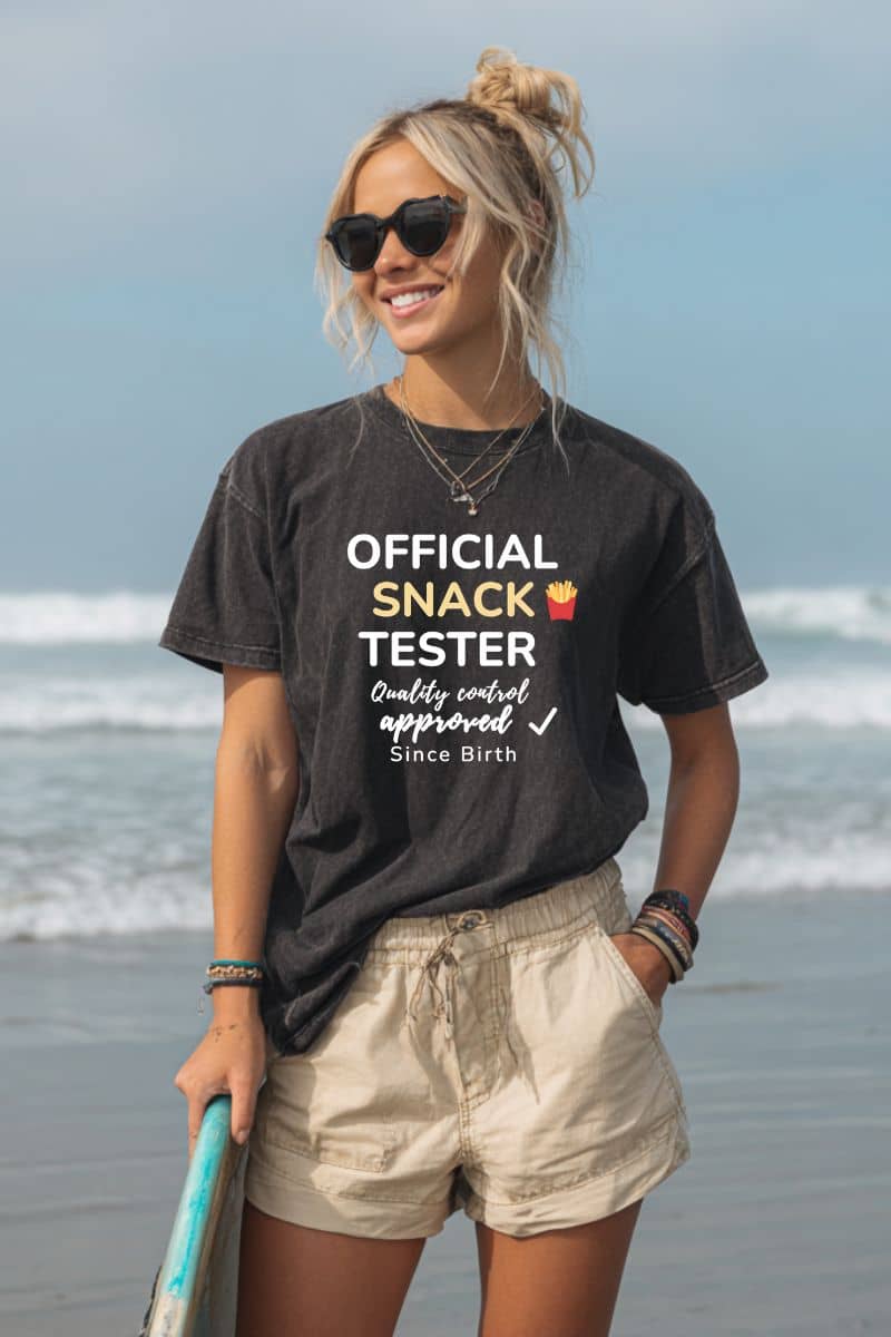 Food shirt with a woman wearing a black t-shirt with text 'Official Snack Tester – Quality Control Approved – Since Birth' on a beach
