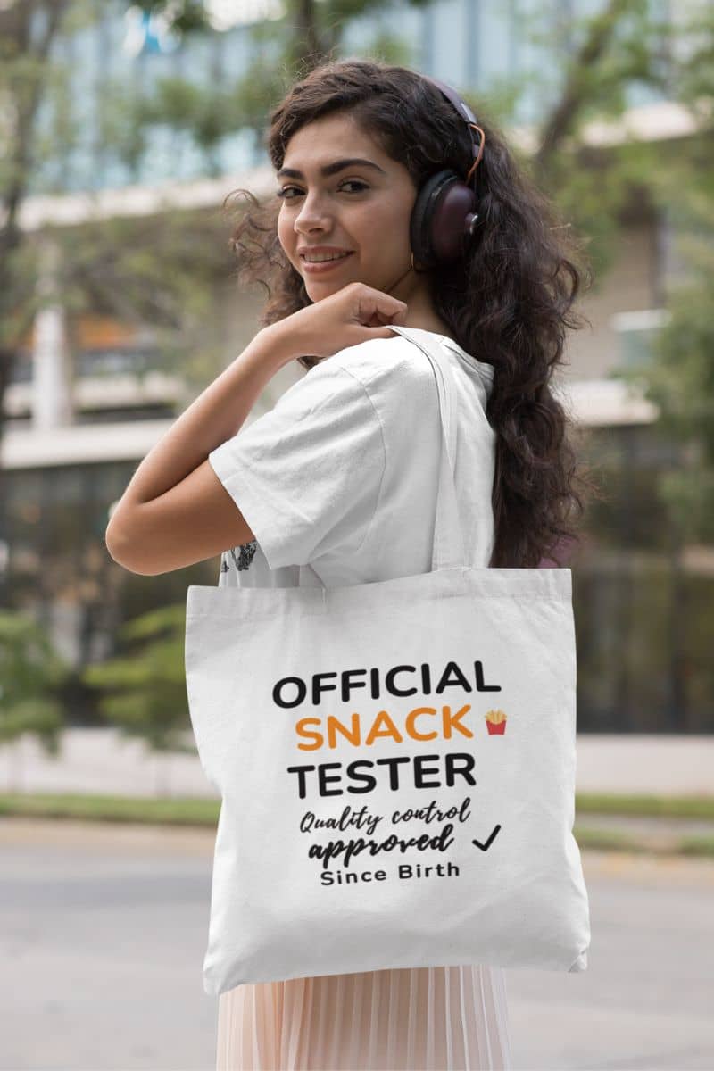 Food tote bag with a woman holding a tote bag with 'Official Snack Tester – Quality Control Approved – Since Birth' text in an urban setting