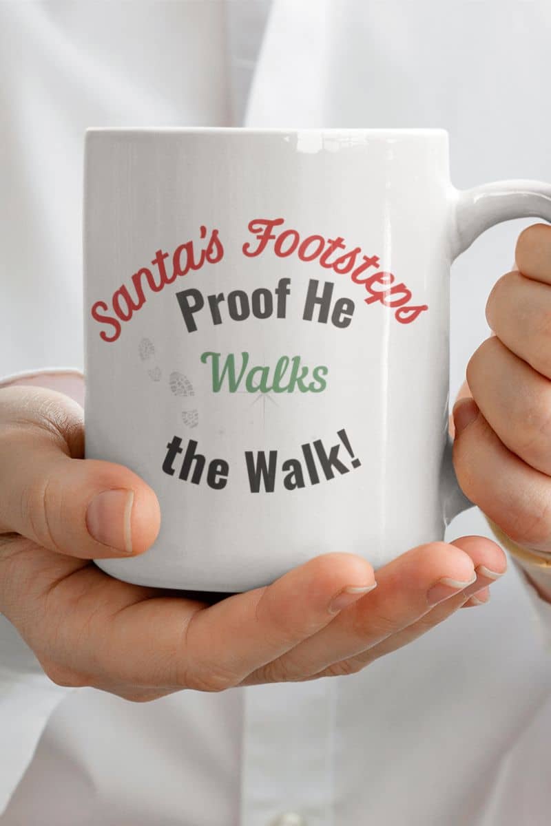 Funny Christmas coffee mug with a white mug with humorous text 'Santa's Footsteps - Proof He Walks The Walk!' held by a person against a white background