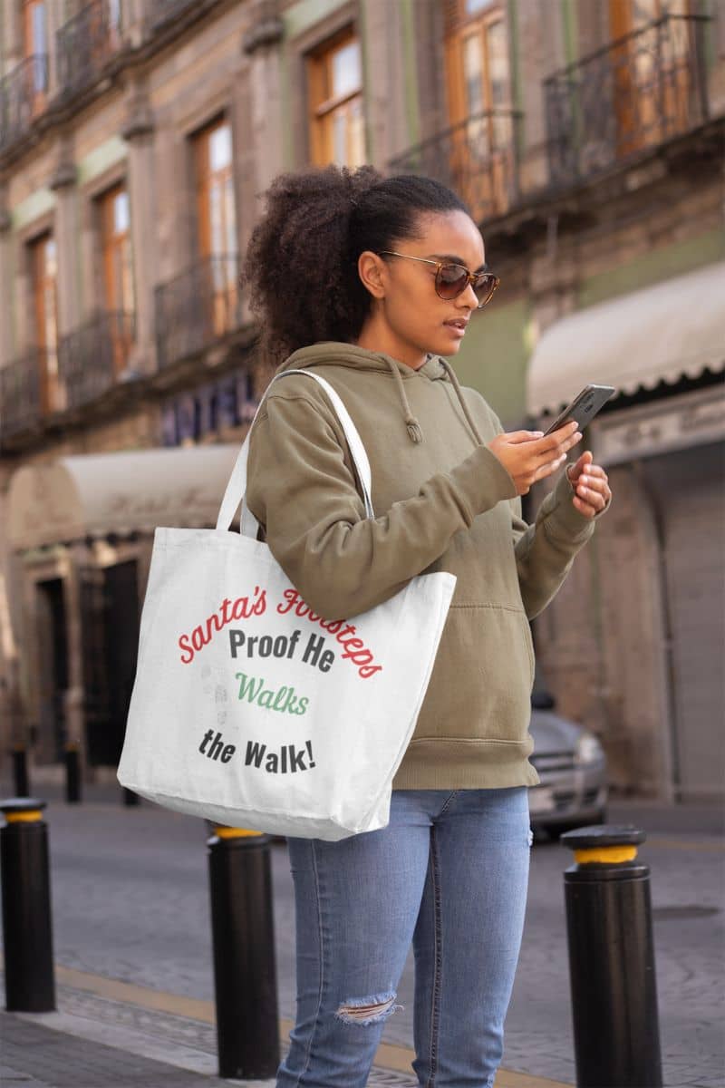 Funny Christmas tote bag with a woman walking on a street holding a phone and a tote bag with text 'Santa's Footsteps - Proof He Walks The Walk!'.