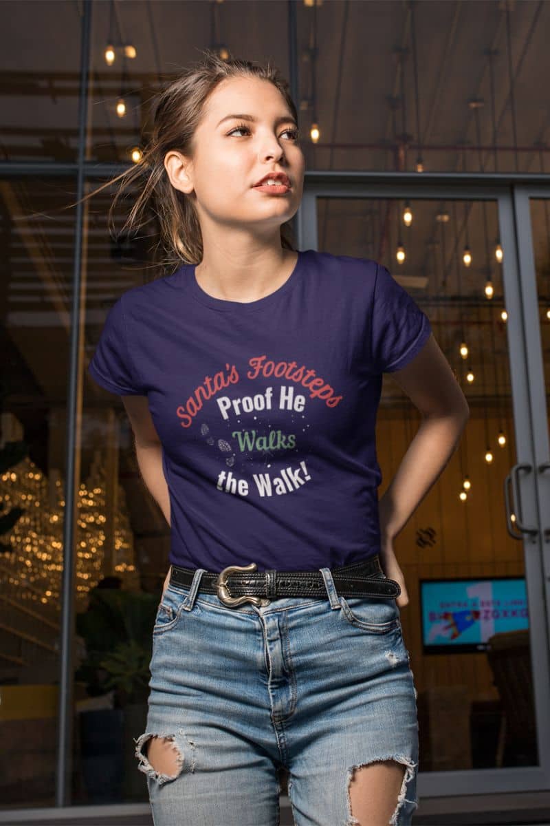 Funny Christmas t-shirt with a woman wearing a purple t-shirt with text 'Santa's Footsteps - Proof He Walks The Walk!', standing in front of a glass door.