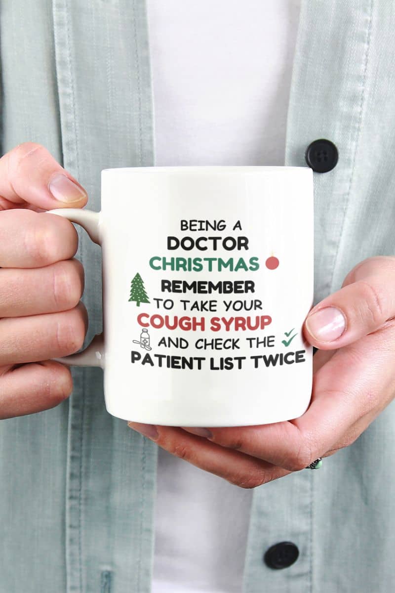 Funny Doctor christmas mug with a person holding a mug with text 'Being a doctor during Christmas, remember to take your cough syrup and check the patient list twice'