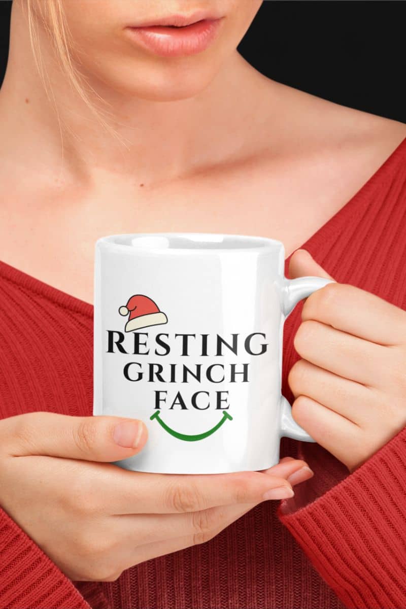 Funny Grinch Christmas mug with a person holding a white mug with 'Resting Grinch Face' text and Santa hat design.