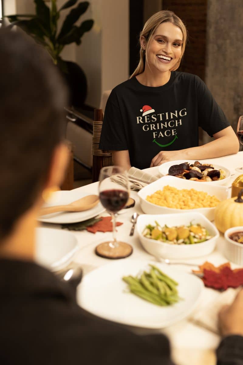 Funny Holiday graphic tee with a person wearing a 'Resting Grinch Face' shirt at a dinner table with food and wine.