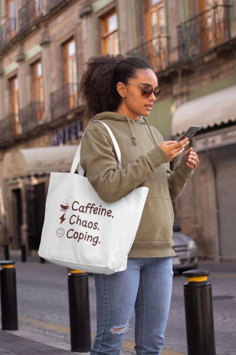 Funny burnout tote with a young woman holding a phone and a tote bag with text  'Caffeine, Chaos & Coping' in an urban setting