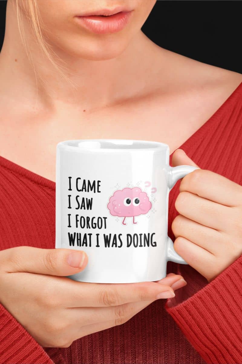 Funny coffee mug with a person holding a white mug with a brain graphic and text 'I came, I saw, I forgot what I was doing', wearing a red sweater.