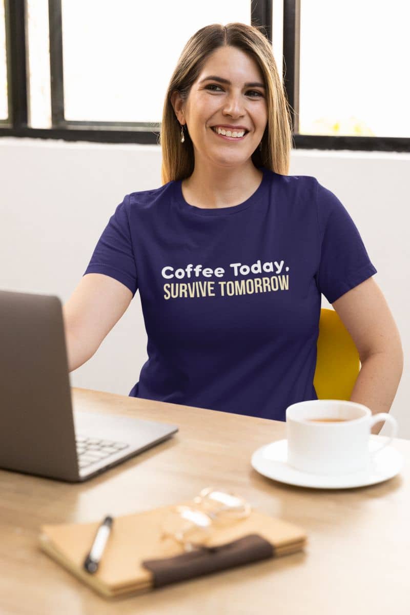 Funny coffee quote shirt with a woman in a purple t-shirt with 'Coffee Today, Survive Tomorrow' text, sitting at a desk with a laptop and coffee.