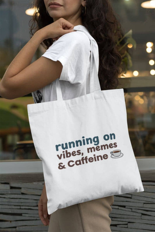 Funny coffee tote bag with a person holding a white tote bag with text 'running on vibes, memes & Caffeine' in an outdoor setting.