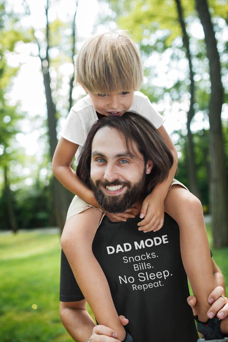Funny dad shirts with a man with a child on his shoulders wearing a black t-shirt with text 'DAD MODE:  Snacks.  Bills. No Sleep. Repeat.' in a park.