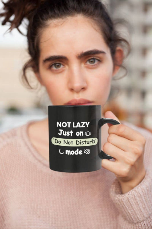 Funny do not disturb mug with a person holding a black mug with text 'Not lazy, Just on 'Do Not Disturb' mode', wearing a light pink sweater.