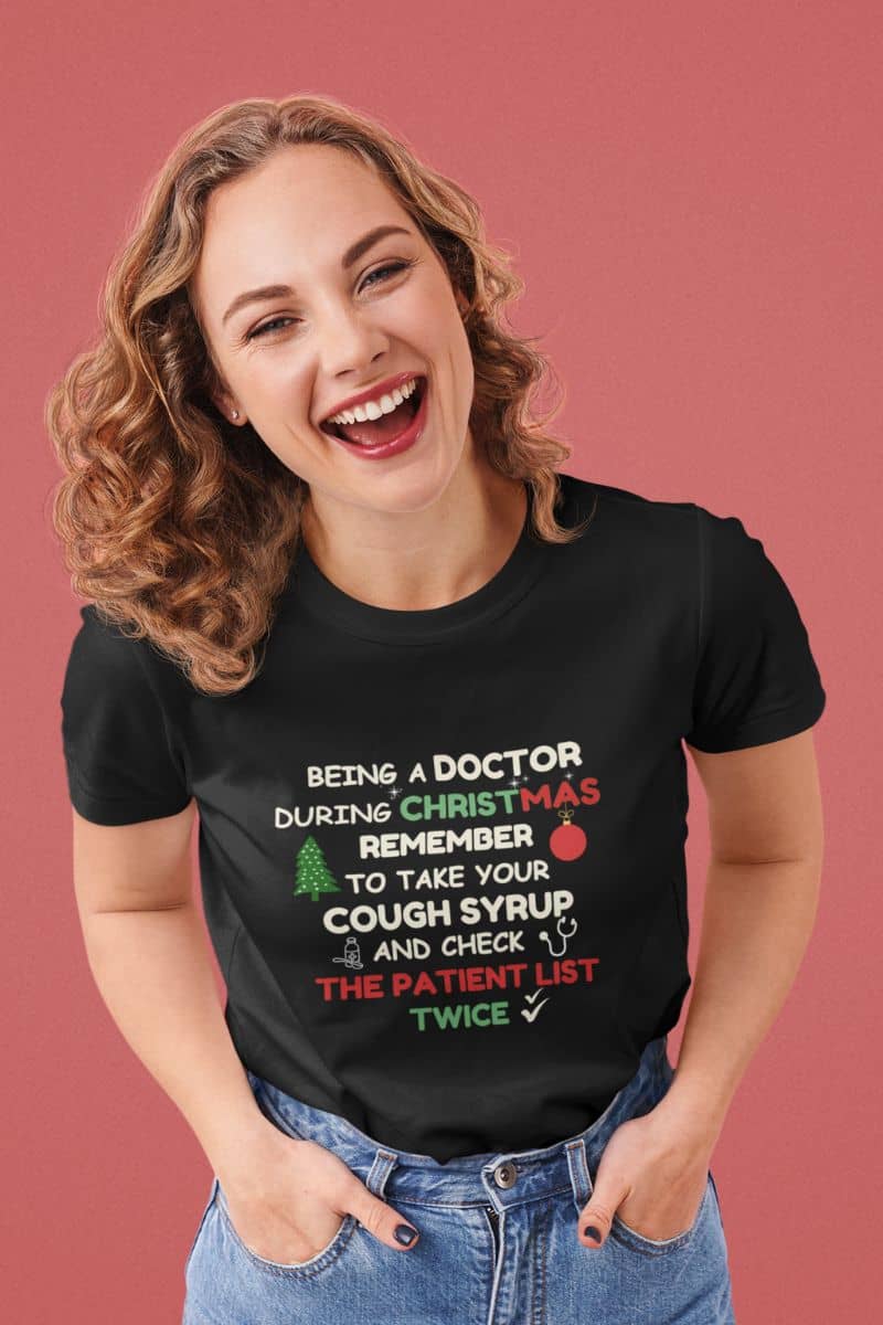 Funny doctor christmas shirt with a woman wearing a black t-shirt with text 'Being a doctor during Christmas, remember to take your cough syrup and check the patient list twice' on a pink background