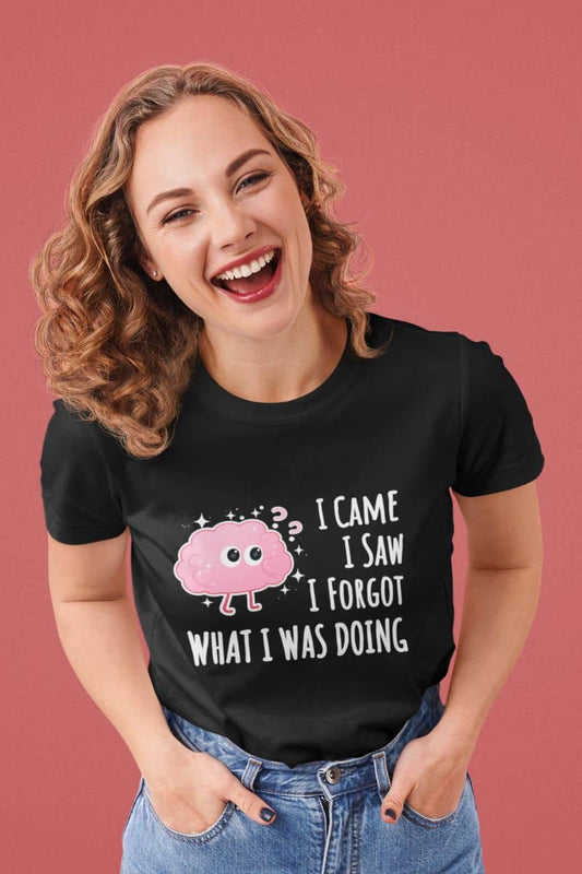 Funny graphic-t-shirt with a woman wearing a black t-shirt with a humorous design and text 'I came, I saw, I forgot what I was doing', laughing against a pink background