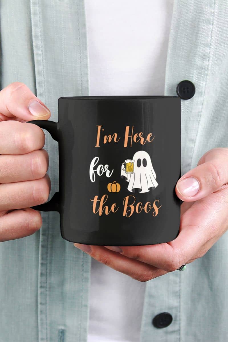 Funny halloween mug with black mug with Halloween design held by a person, featuring text 'I’m Here for the Boos'