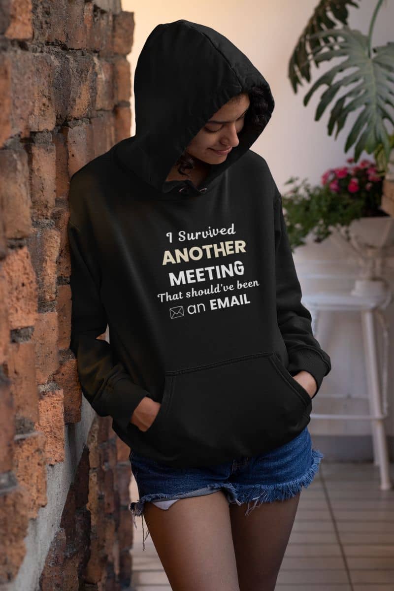 Funny hoodie with a person wearing a black hoodie with text 'I Survived Another Meeting That Should’ve Been an Email', standing against a brick wall.