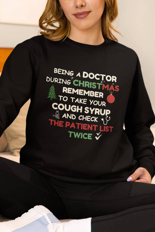 Funny medical christmas sweatshirt with a person in a black sweatshirt featuring the text 'Being a doctor during Christmas, remember to take your cough syrup and check the patient list twice'