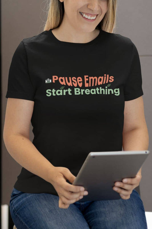 Funny meditation t-shirt for work with a person wearing a black t-shirt with text 'Pause Emails Start Breathing', holding a tablet.