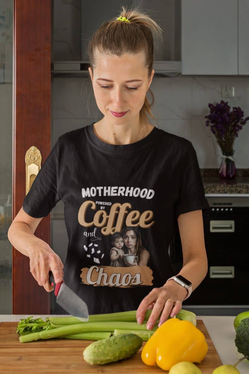Funny mom shirt with a woman in a kitchen wearing a black t-shirt with text 'Motherhood: Powered by Coffee and Chaos.', cutting vegetables.