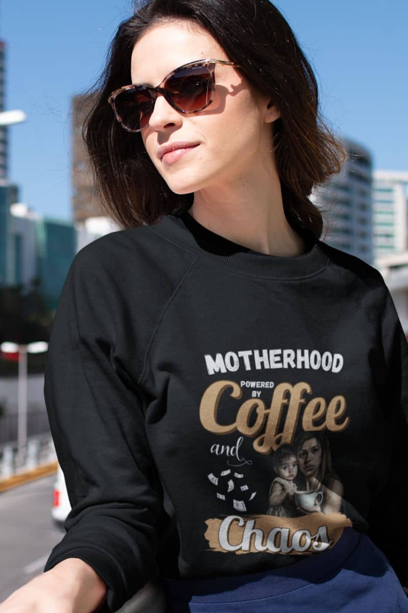 Funny mom sweatshirt with a woman wearing a black sweatshirt with text 'Motherhood powered by Coffee and Chaos' and graphics, standing outdoors.